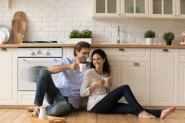 Jeune couple tient tasses assis sur le sol en bois dans la cuisine — Photo