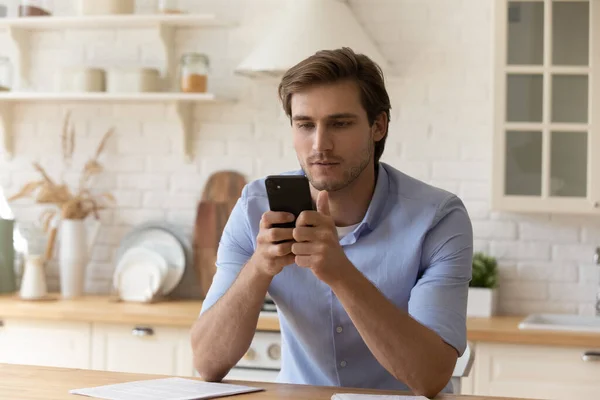Grave bell'uomo d'affari siede in cucina domestica con smartphone in mano — Foto Stock