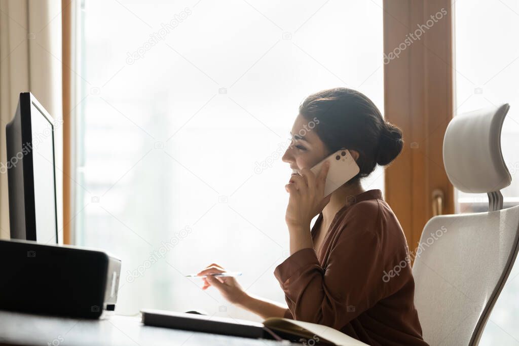 Smiling Indian business lady sit at workplace talk on smartphone