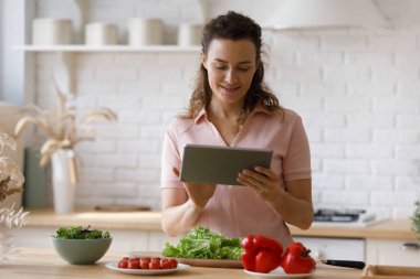 Mutlu genç kadın dijital tablet kullanıyor, evde yemek pişiriyor..