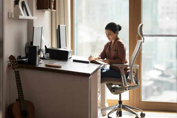 Serious Indian female sit at desk holds pen jotting information — Foto de Stock