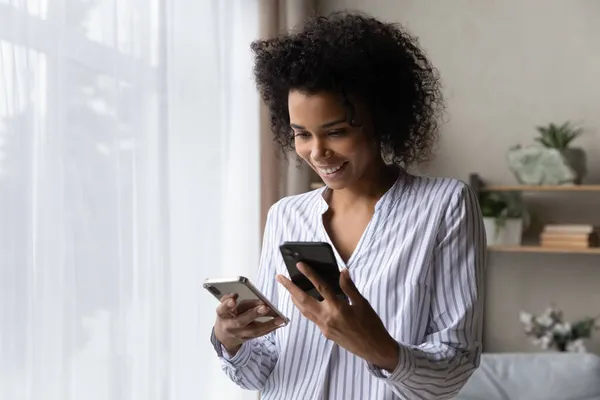 Leende afroamerikansk kvinna med hjälp av två smartphones, står hemma — Stockfoto