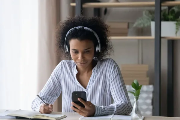 Focused African American woman in headphones taking notes, holding smartphone — Stock Photo, Image
