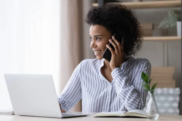 Smiling African American businesswoman using laptop and talking on phone — Stock Photo, Image