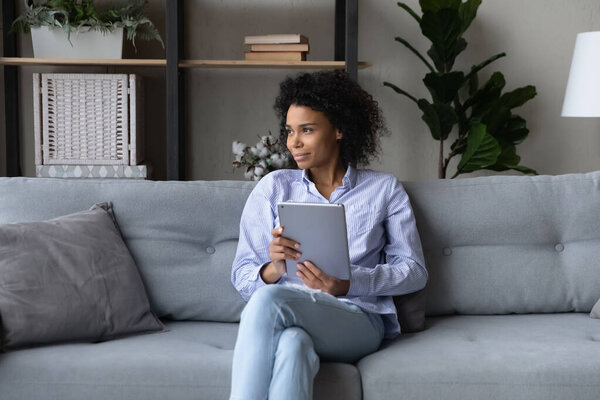 Dreamy thoughtful African American woman looking in distance, holding tablet