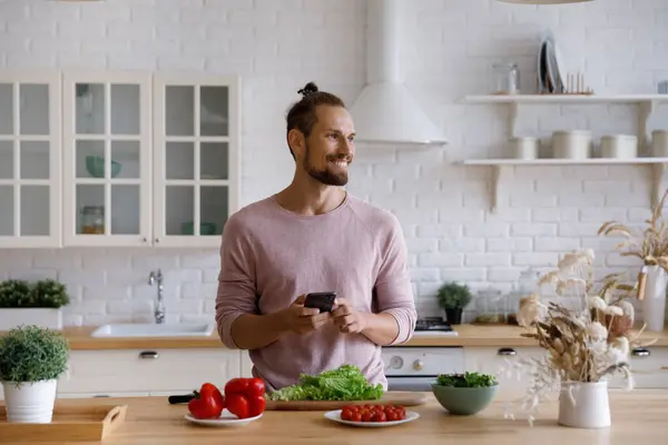 Distrait de la cuisson des aliments heureux jeune homme en utilisant un smartphone. — Photo