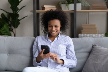 Akıllı telefonuyla kanepede dinlenen gülümseyen Afrikalı Amerikalı kadın.