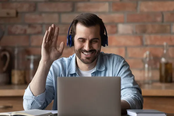Positive young male in headphones wave hand to laptop screen — Stock Photo, Image