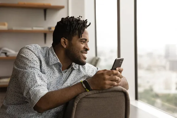 Sorrindo mestiço jovem com celular sentindo-se sonhador. — Fotografia de Stock