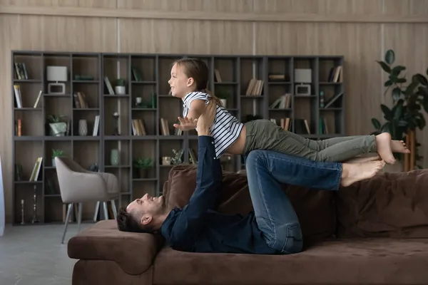 Sonriente padre amoroso levantando a su hijita fingiendo volar en el sofá —  Fotos de Stock