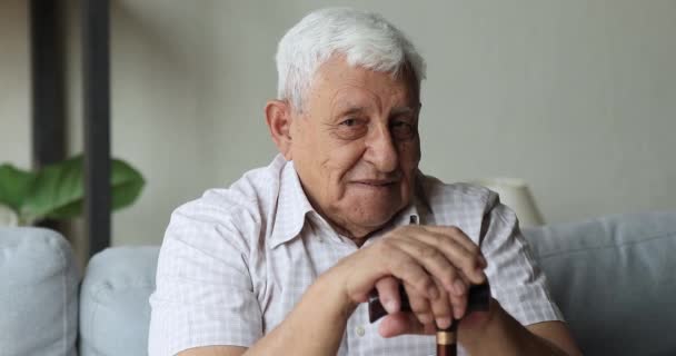Optimistický starší muž se zdravotním postižením sedí na pohovce držet hůl — Stock video