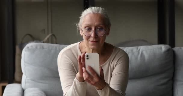 Стримана жінка в окулярах сидить на дивані, використовуючи сучасний смартфон — стокове відео