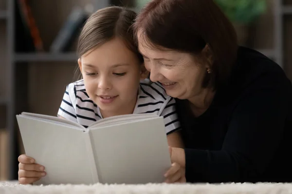 Улыбающаяся взрослая бабушка с маленькой девочкой, читающей вместе книгу — стоковое фото