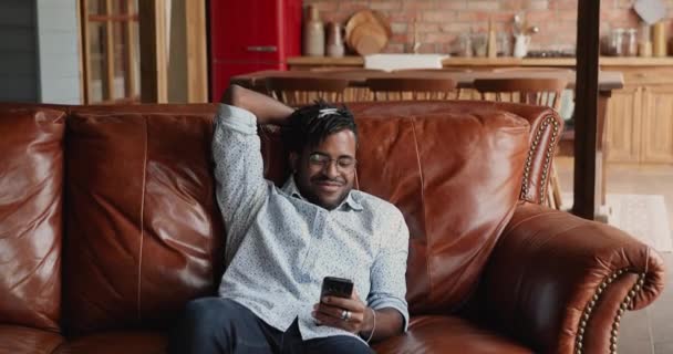 Afrikaner entspannt sich auf Sofa mit Handy und schaut sich Online-Videos an — Stockvideo