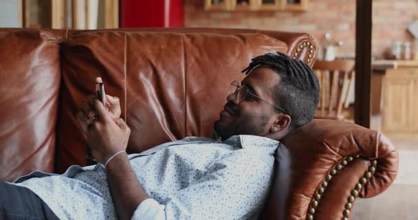 リラックスしたアフリカ人男性横たわっソファに自宅でスマートフォン — ストック動画
