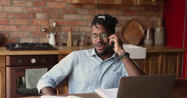 Afrikaanse man zit aan tafel met laptop praten over de telefoon — Stockvideo