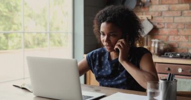 Akıllı telefondan konuşan çekici Afrikalı kadın evde dizüstü bilgisayar kullanıyor.