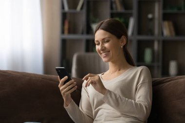 Gülümseyen güzel kadın kanepeye oturur ve akıllı telefon ekranına bakar.