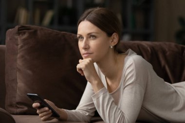 Genç ciddi bir kadın akıllı telefonuyla kanepede uzanmış dalgın bakıyor.