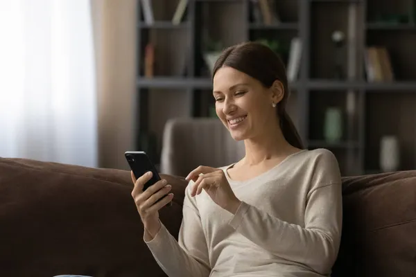Leende vacker kvinna sitta på soffan tittar på smartphone skärm — Stockfoto