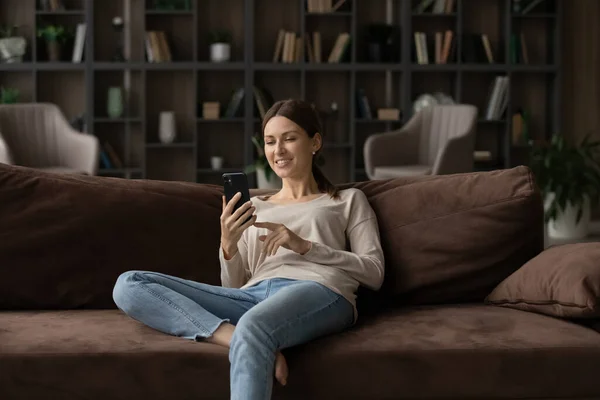Jovem mulher passar lazer detém smartphone enquanto relaxa em casa — Fotografia de Stock