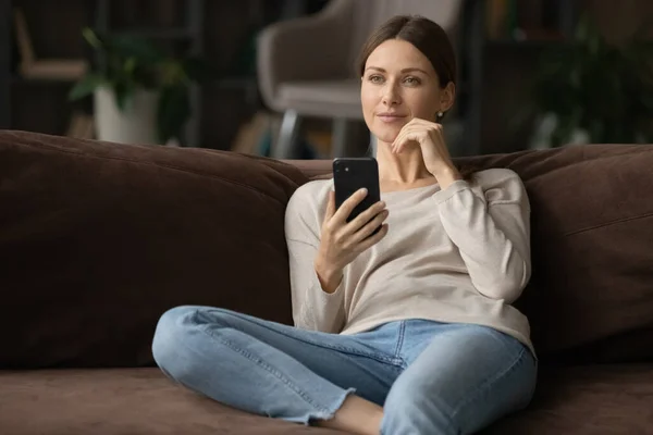 Mladá žena sedí na pohovce drží smartphone vypadá zamyšleně — Stock fotografie
