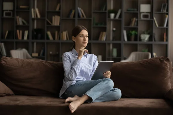Mulher pensativa senta-se no sofá com tablet digital — Fotografia de Stock