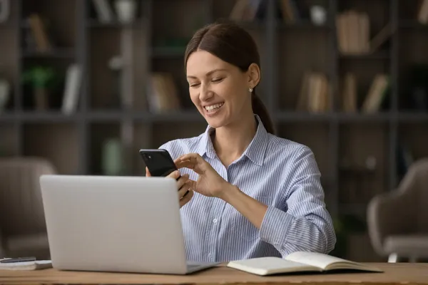 Šťastná obchodní žena sedět na pracovišti v blízkosti notebooku drží smartphone — Stock fotografie