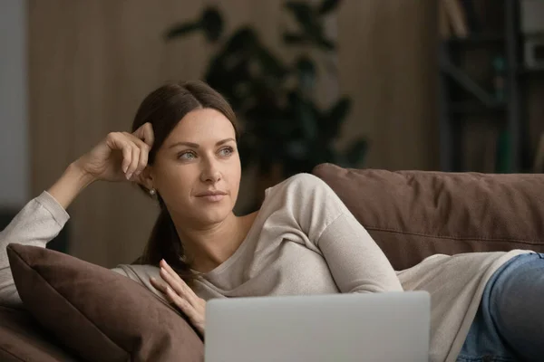 Donna sognante sdraiata sul divano con laptop guardando in lontananza — Foto Stock