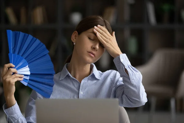 Unhealthy overheated woman sit at workplace waving with hand fan — Stock Photo, Image