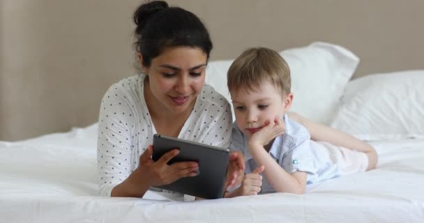 Индийская мать и маленький кавказский сын используют планшет на кровати — стоковое видео