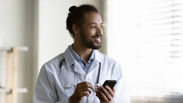 Sonriente médico afroamericano sosteniendo teléfono inteligente, mirando a la distancia —  Fotos de Stock