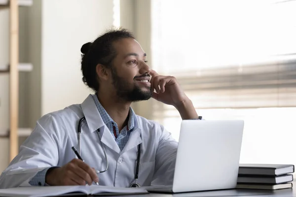 Pensativo sonriente hombre afroamericano médico tomando notas, utilizando el ordenador portátil — Foto de Stock