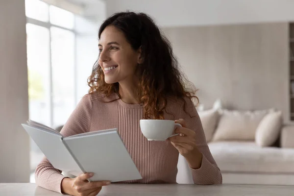 Soñadora mujer sonriente sosteniendo libro y taza de té visualizando — Foto de Stock