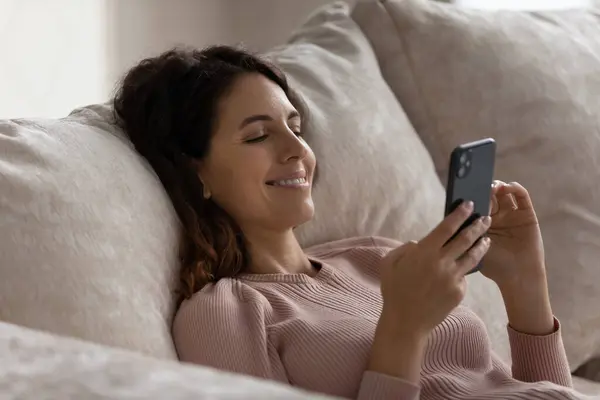 Cerca de la mujer sonriente relajarse con el teléfono inteligente en el sofá cómodo — Foto de Stock