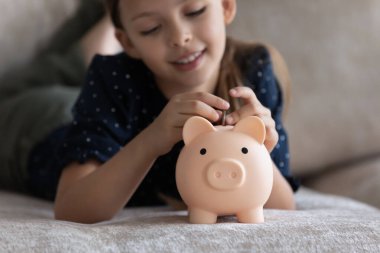 Little girl lying on sofa put coin into piggy bank clipart