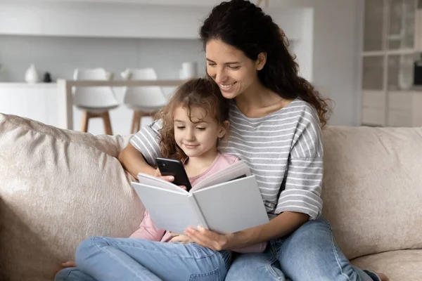 Sonriente madre con hija distraída del libro, adicción a los dispositivos —  Fotos de Stock