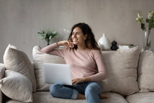 Happy dreamy woman with laptop sitting on cozy couch