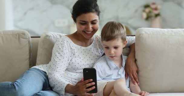 Šťastná indická matka objetí malý syn na gauči držet smartphone — Stock video