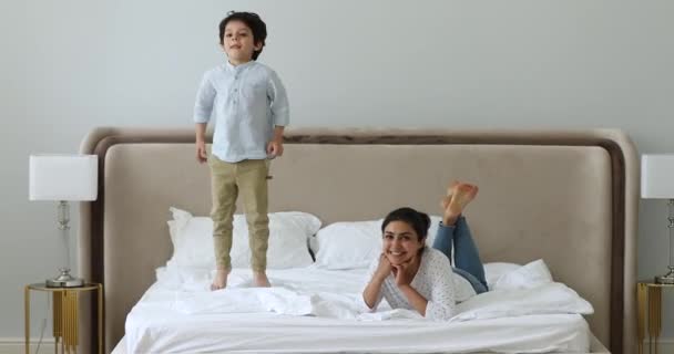 Улыбающаяся индийская мамочка расслабляется, пока маленький сын прыгает на кровать — стоковое видео