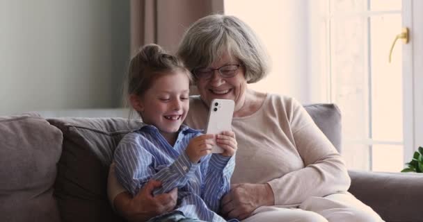 Liebevolle alte Oma beobachtet kleine Enkelin beim Spielen am Telefon — Stockvideo