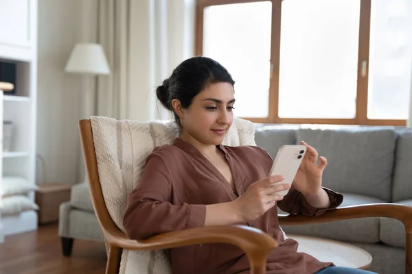 Avslappnad ung indian kvinna med mobiltelefon, sitter i fåtölj. — Stockfoto