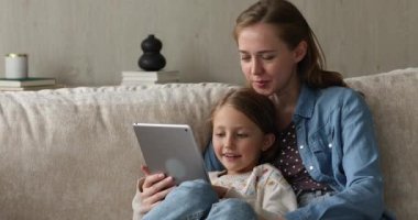 Genç anne, küçük kızıyla birlikte dokunmatik yastıkta kitap okuyor.