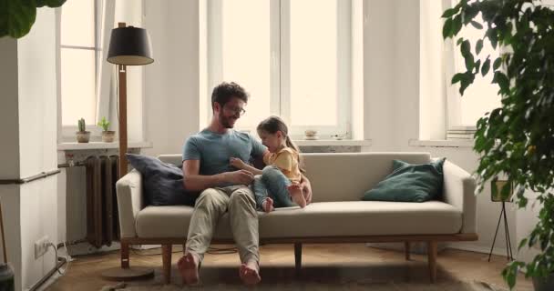 Fiatal nevelőapa csiklandozik a kanapén a kis örökbefogadott lánnyal. — Stock videók