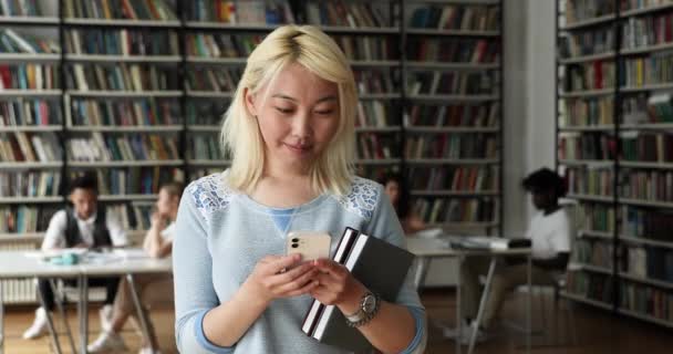 Asian student girl standing in library holds cellphone texting message — Stock Video