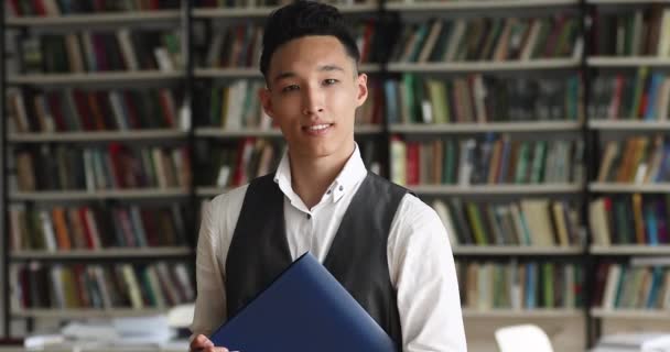 Asiático chico celebración carpeta mirar en cámara pose en biblioteca — Vídeo de stock
