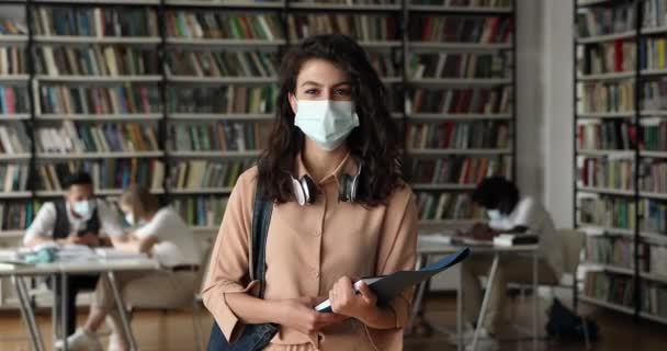 Muchacha atractiva estudiante usar máscara facial protectora posando en la biblioteca — Vídeo de stock