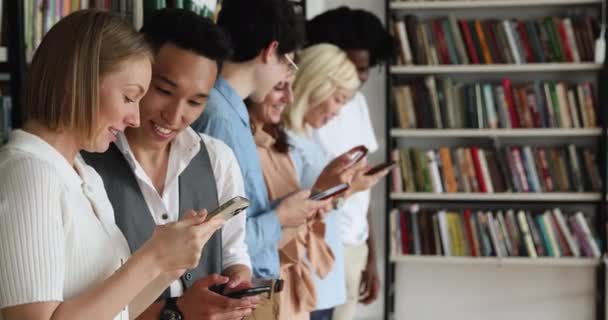 Diversos estudiantes en fila en la biblioteca con teléfonos inteligentes — Vídeo de stock