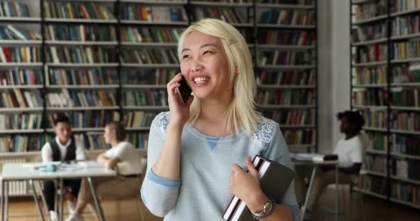 Ελκυστική Ασιάτισσα φοιτήτρια στέκεται στη βιβλιοθήκη φλυαρία στο τηλέφωνο — Αρχείο Βίντεο