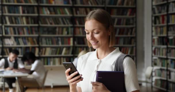 Junge attraktive Studentin steht mit Handy in öffentlicher Bibliothek — Stockvideo